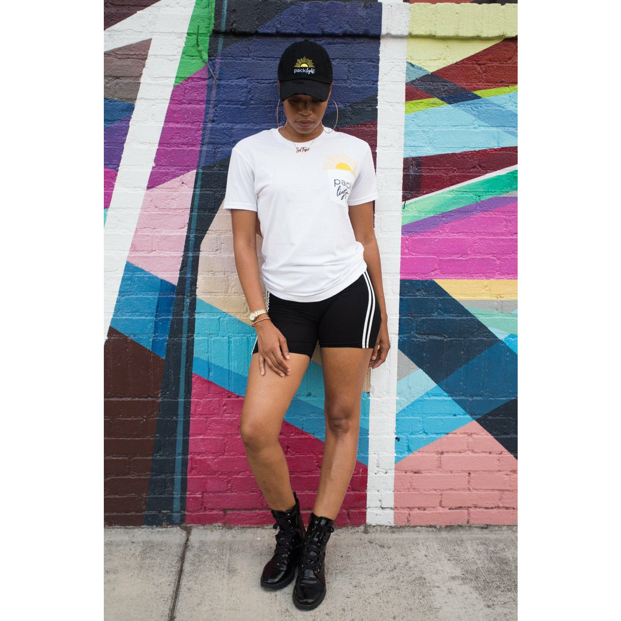 woman wear Pack Light hat and shirt standing in front of vibrant mural