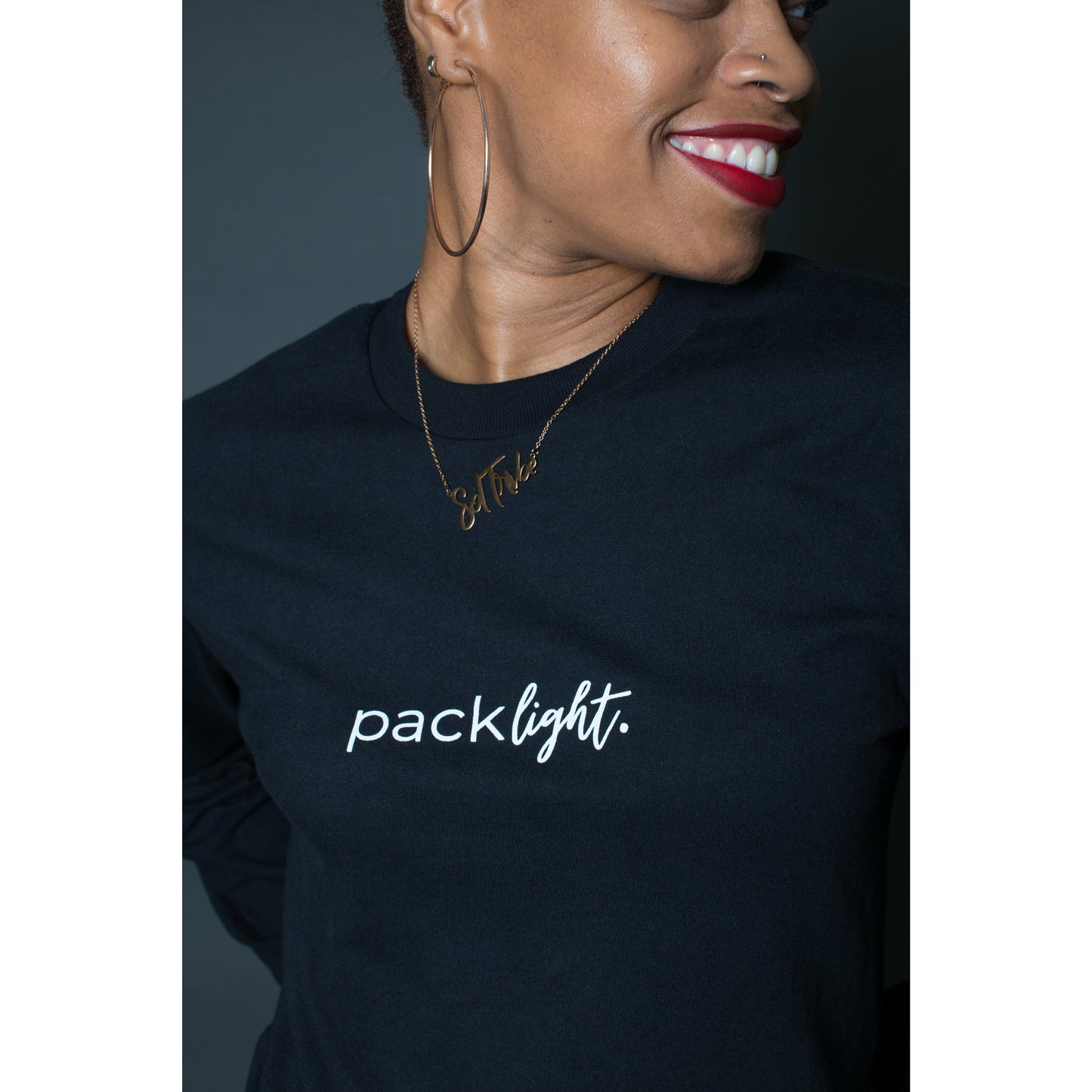 smiling woman model wearing black affirmation tee that says Pack Light on the sleeve and front of the tee by Sol Rise Essentials